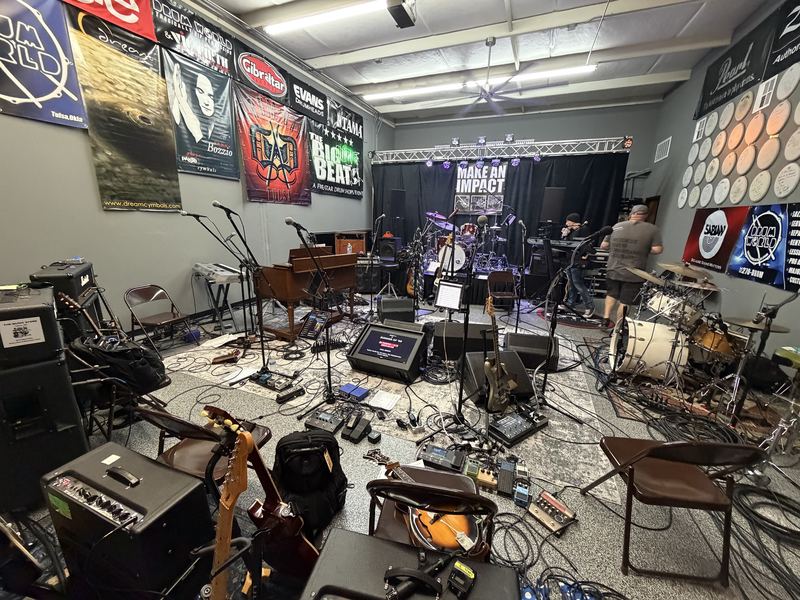 Stage setup in a rehearsal space with drum kit, guitar amps, keyboards and cabling