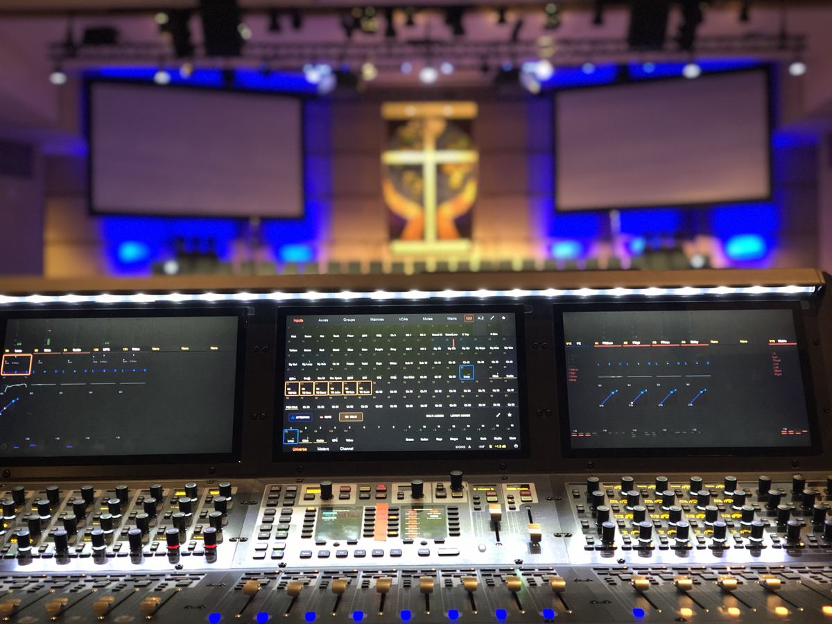 Digital mixing console with three touchscreens at front of house in a church sanctuary, with two projection screens, a lit cross and blue stage lighting behind