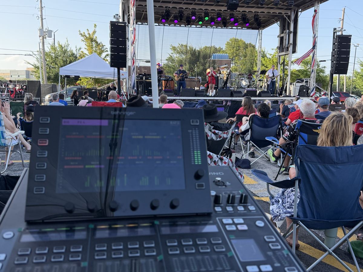 Digital mixing console at front of house facing an outdoor festival stage with a crowd in lawn chairs