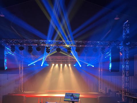 Blue moving-head beams fanning up through haze from a truss rig over a stage, with warm wash light on the stage deck