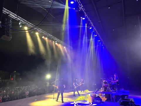 Band performing on an outdoor stage at night under blue and yellow beams from a side truss, with line-array speakers overhead and a crowd at the edge