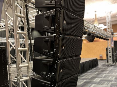 Side view of a line array of RCF speaker cabinets hung beside a truss tower in a ballroom, with a lighting truss, skirted stage and second truss tower behind