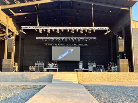Covered outdoor stage with truss lighting rigs, a video screen, speaker stacks at each side and a walkway leading to the stage